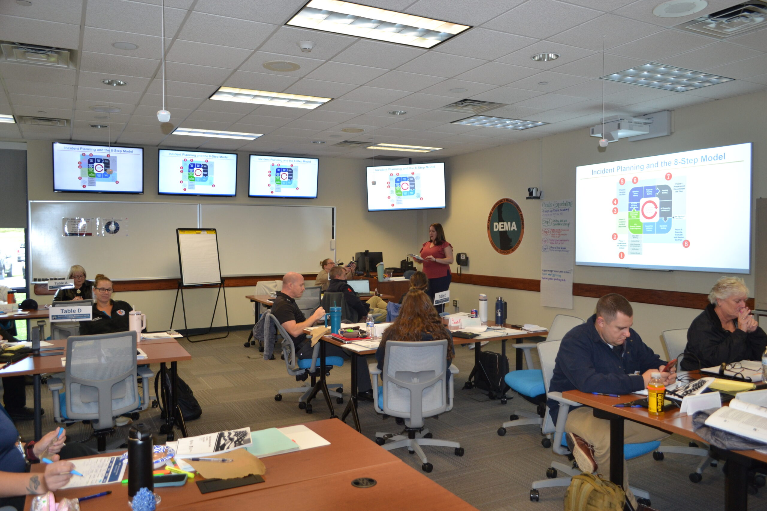 A classroom setting with students seated at tables facing multiple screens displaying a presentation slide, while an instructor stands near a whiteboard.