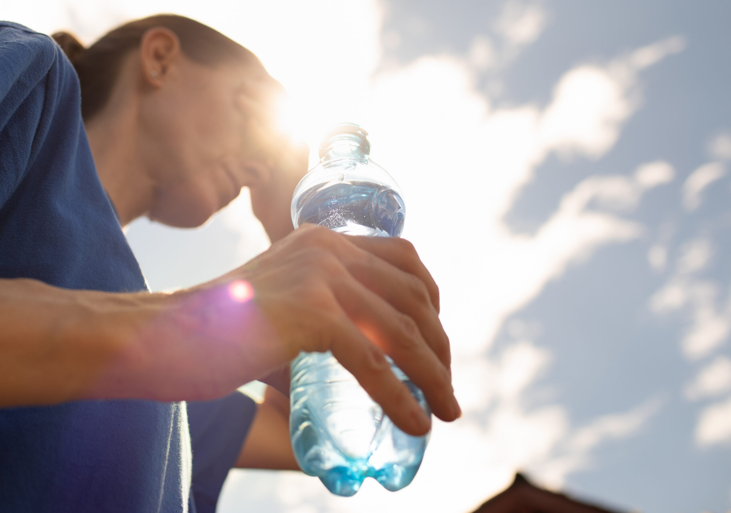 Person holding a clear plastic water bottle against a bright sky with sunlight shining through